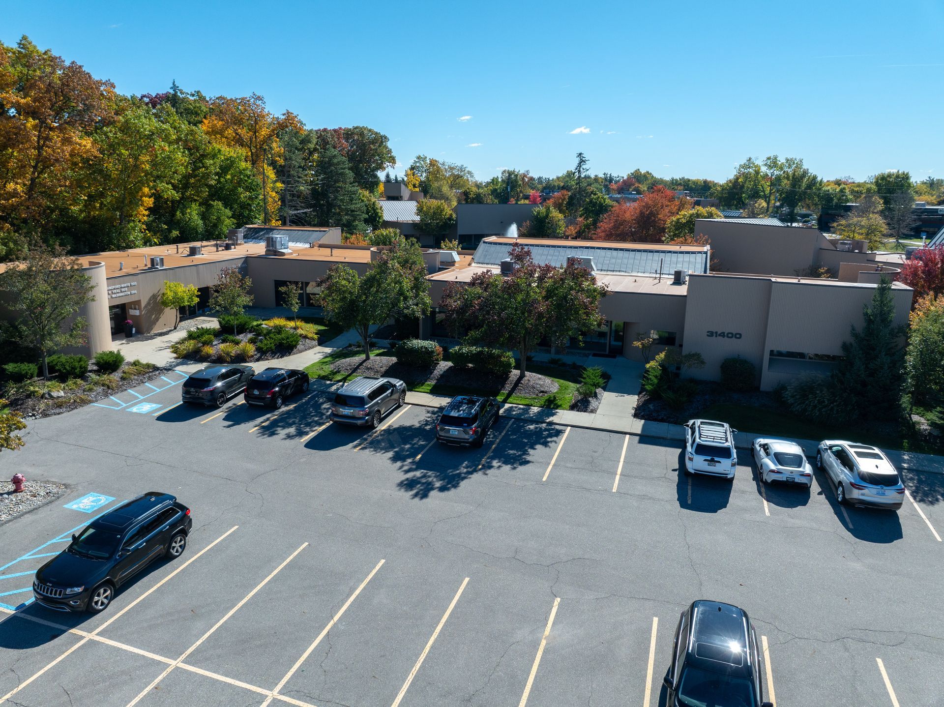 Aerial view of building with parking lot and cars under a blue sky, surrounded by trees.