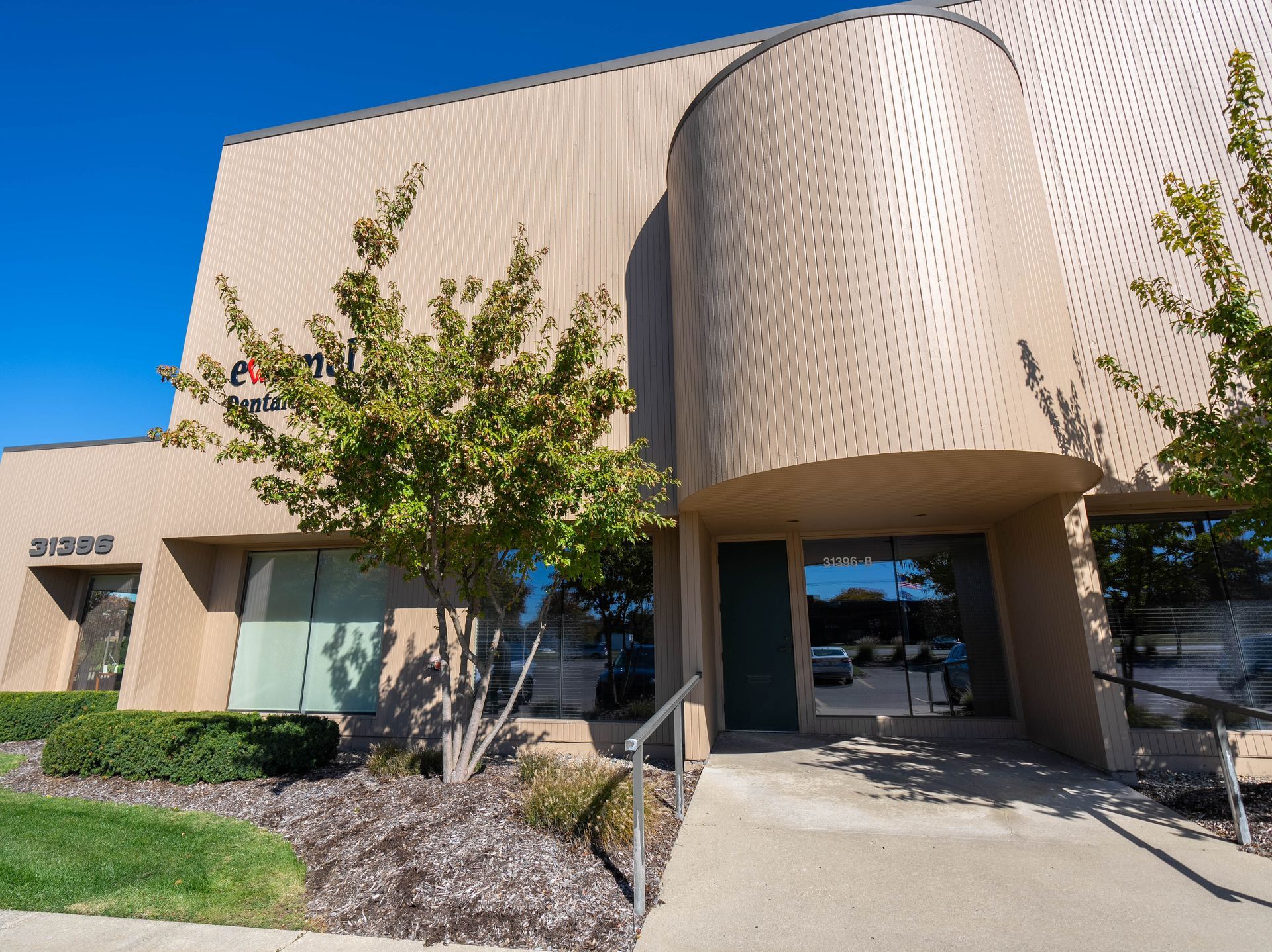 Tan office building with a curved, perforated facade; green trees, blue sky.