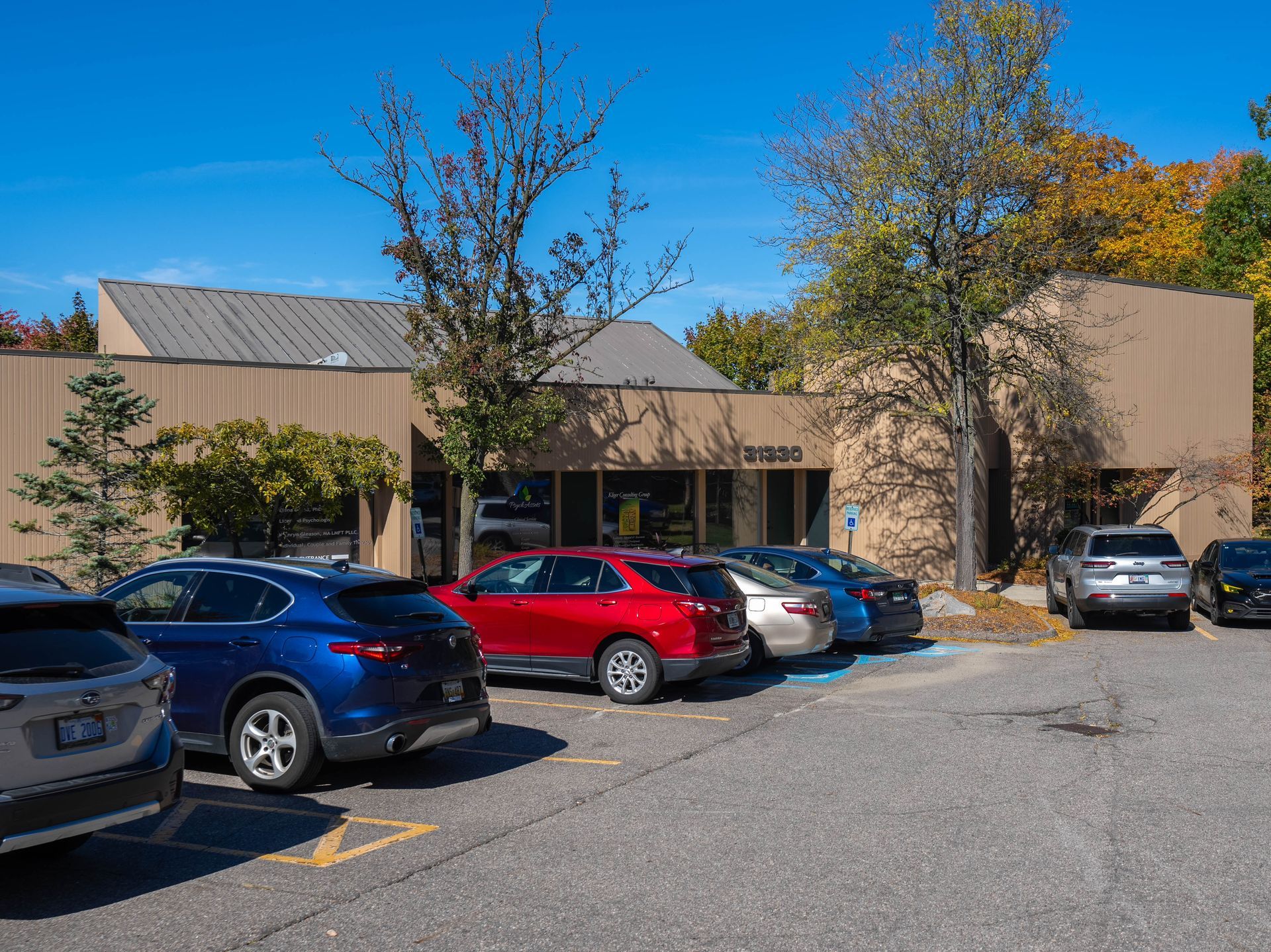 Cars parked in front of a one-story tan building with a blue sky and fall trees.