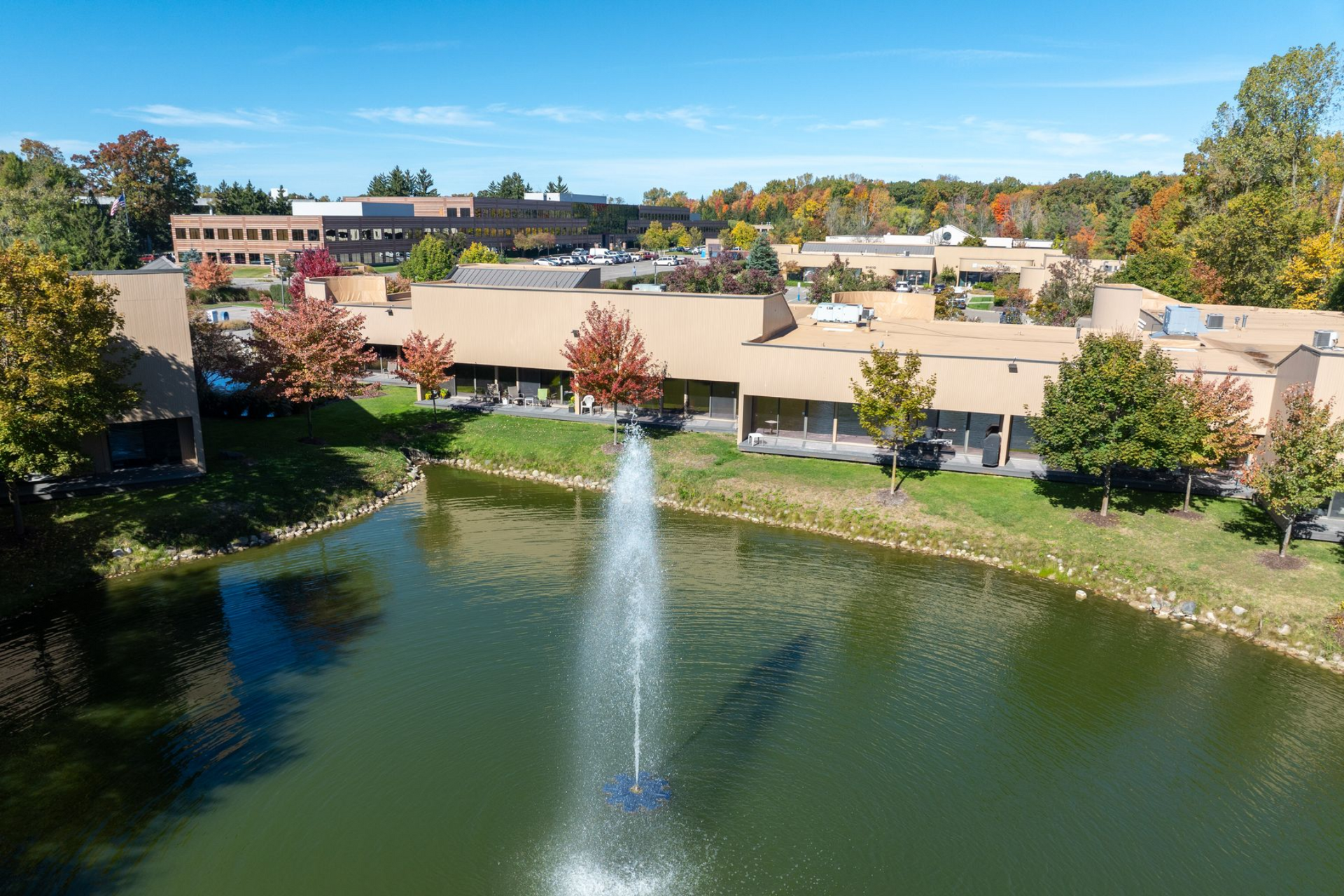 Pond with fountain in front of a low-rise building and trees under a blue sky.