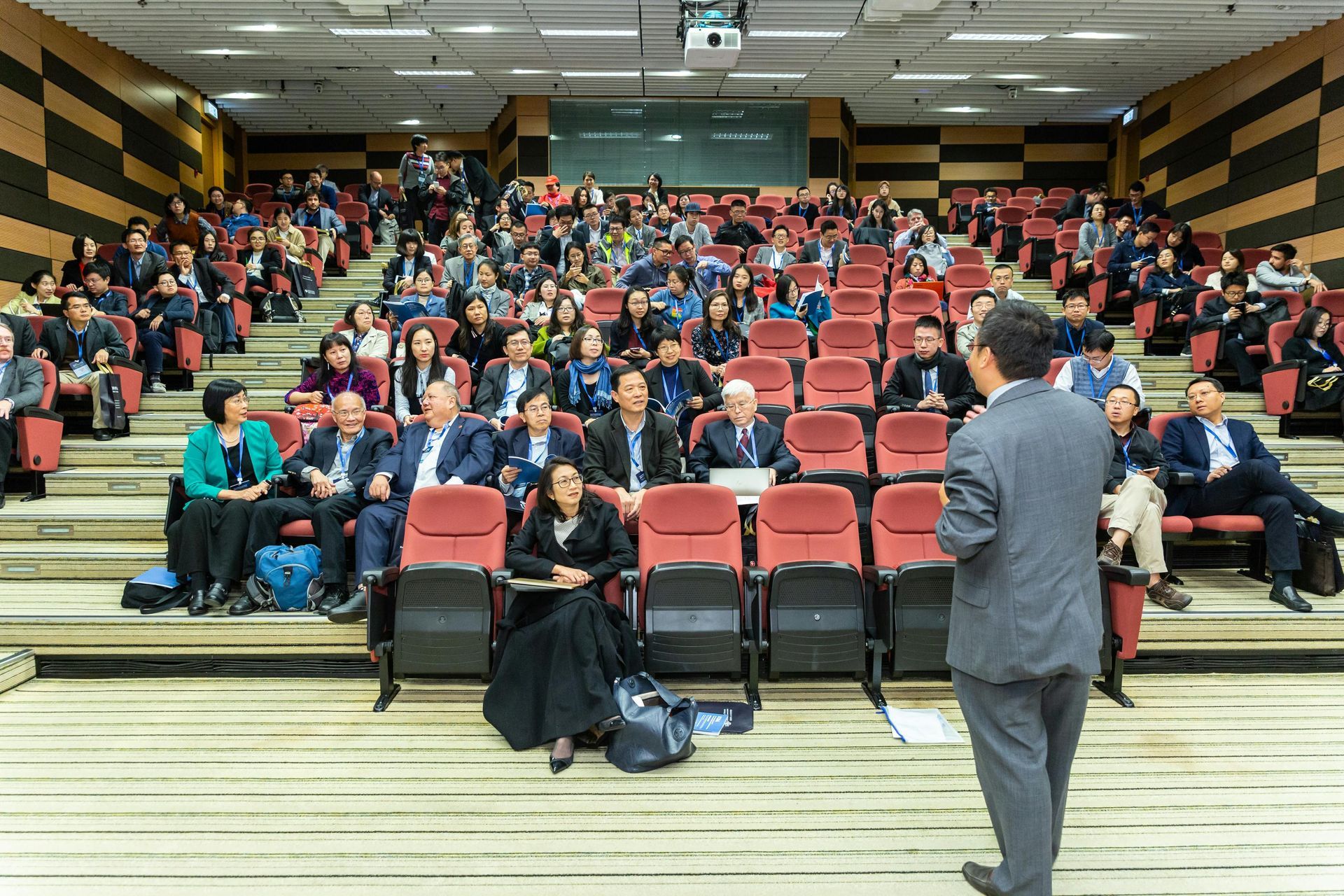 A speaker stands before a large audience in a tiered lecture hall with red seats and wood-paneled walls.