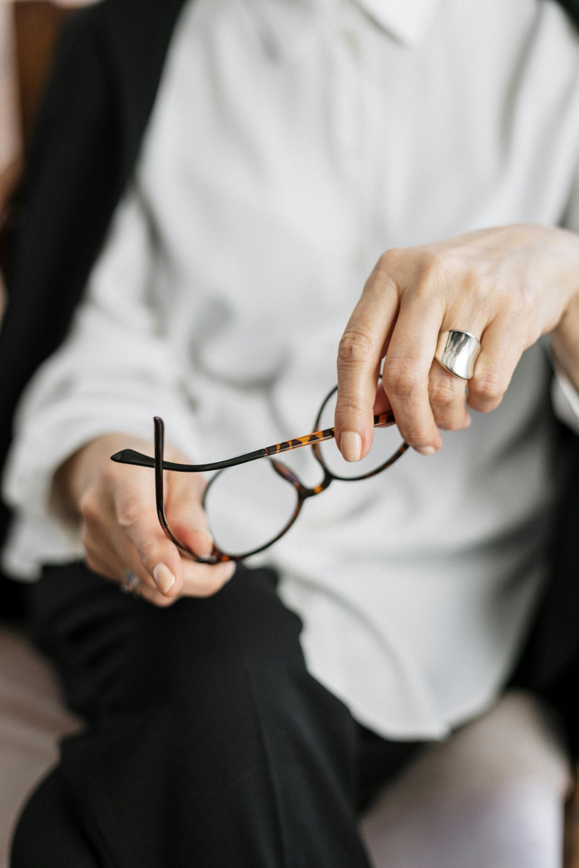 Hands holding a pair of eyeglasses, wearing a silver ring, with a light shirt and dark jacket in the background.