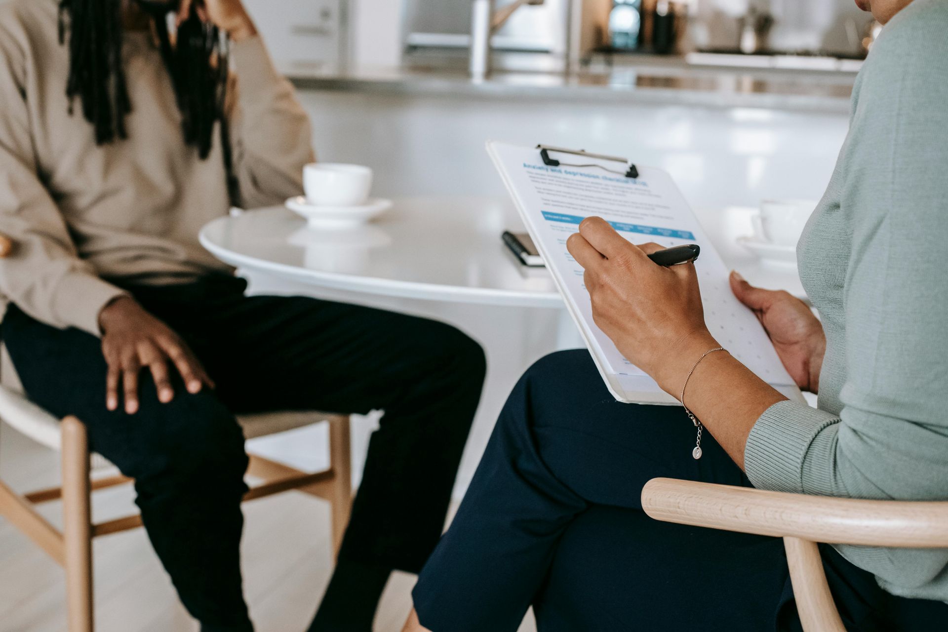 A professional sits at a table with a person, holding a clipboard and pen to record information during a consultation.