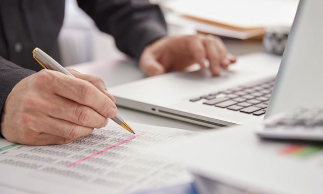 A person is writing on a piece of paper in front of a laptop computer.