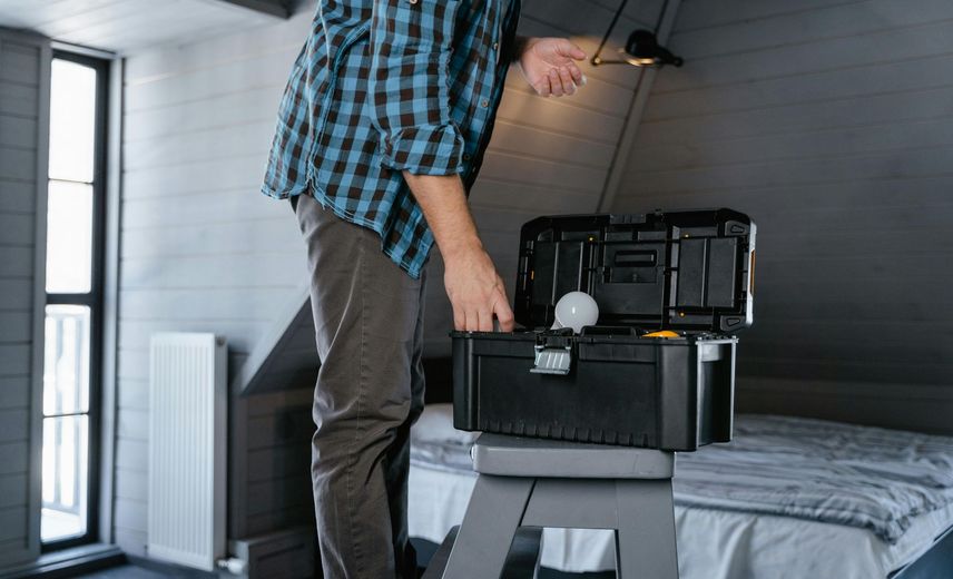 Person in plaid shirt opening a black toolbox on a small platform in a room.