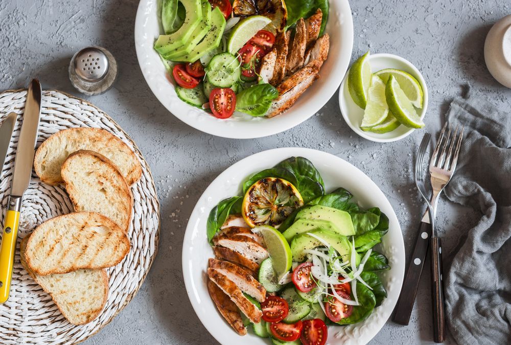 Two bowls of salad with chicken , avocado , tomatoes and cucumber on a table.