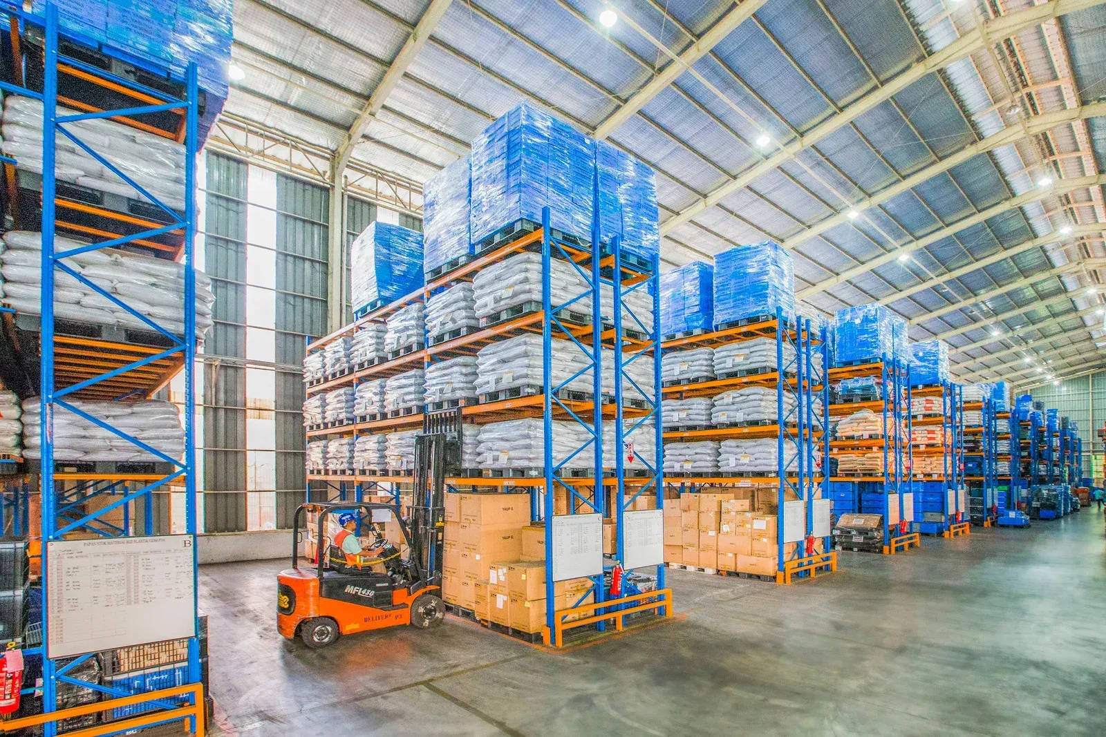 Warehouse interior with forklift loading shelves of boxes and wrapped pallets.