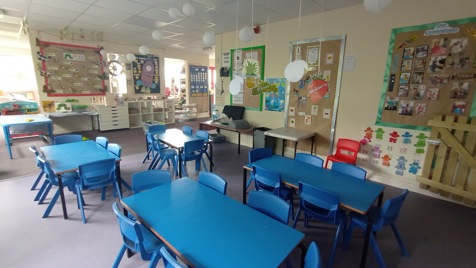 A classroom with blue tables and chairs and a bulletin board Gingerbread