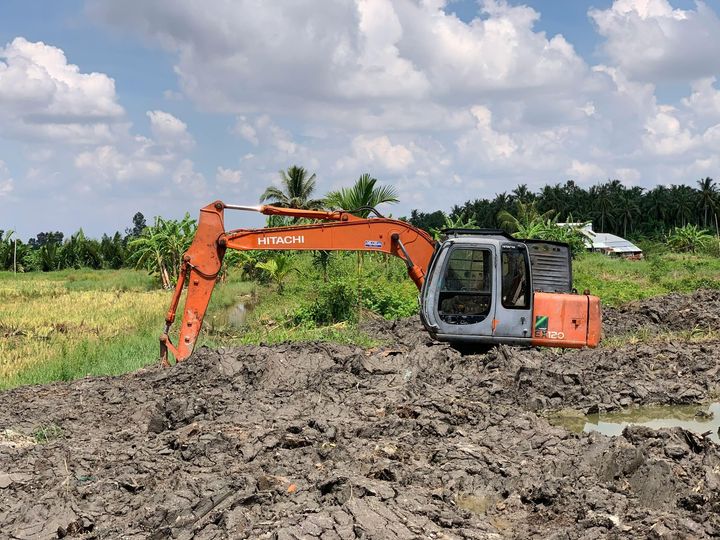 An orange Hitachi excavator sits idle on muddy, churned-up ground in an open field under a blue, cloudy sky.