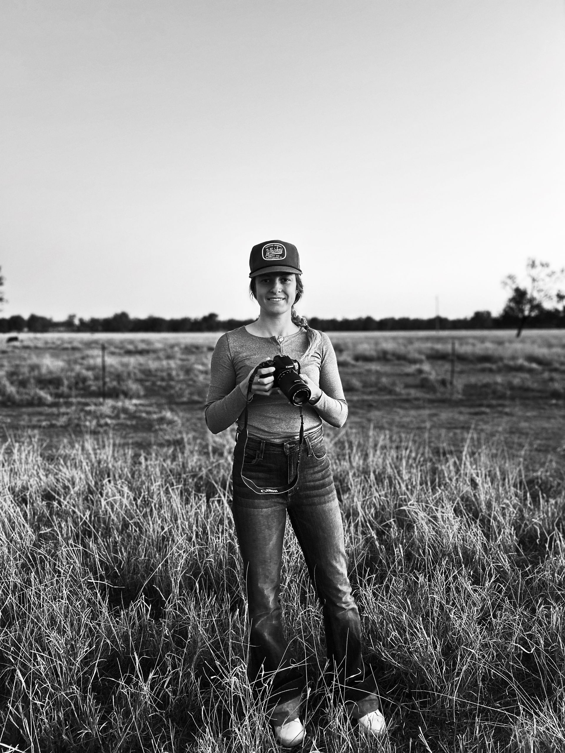 Person in a field, holding binoculars, wearing a cap and jeans.