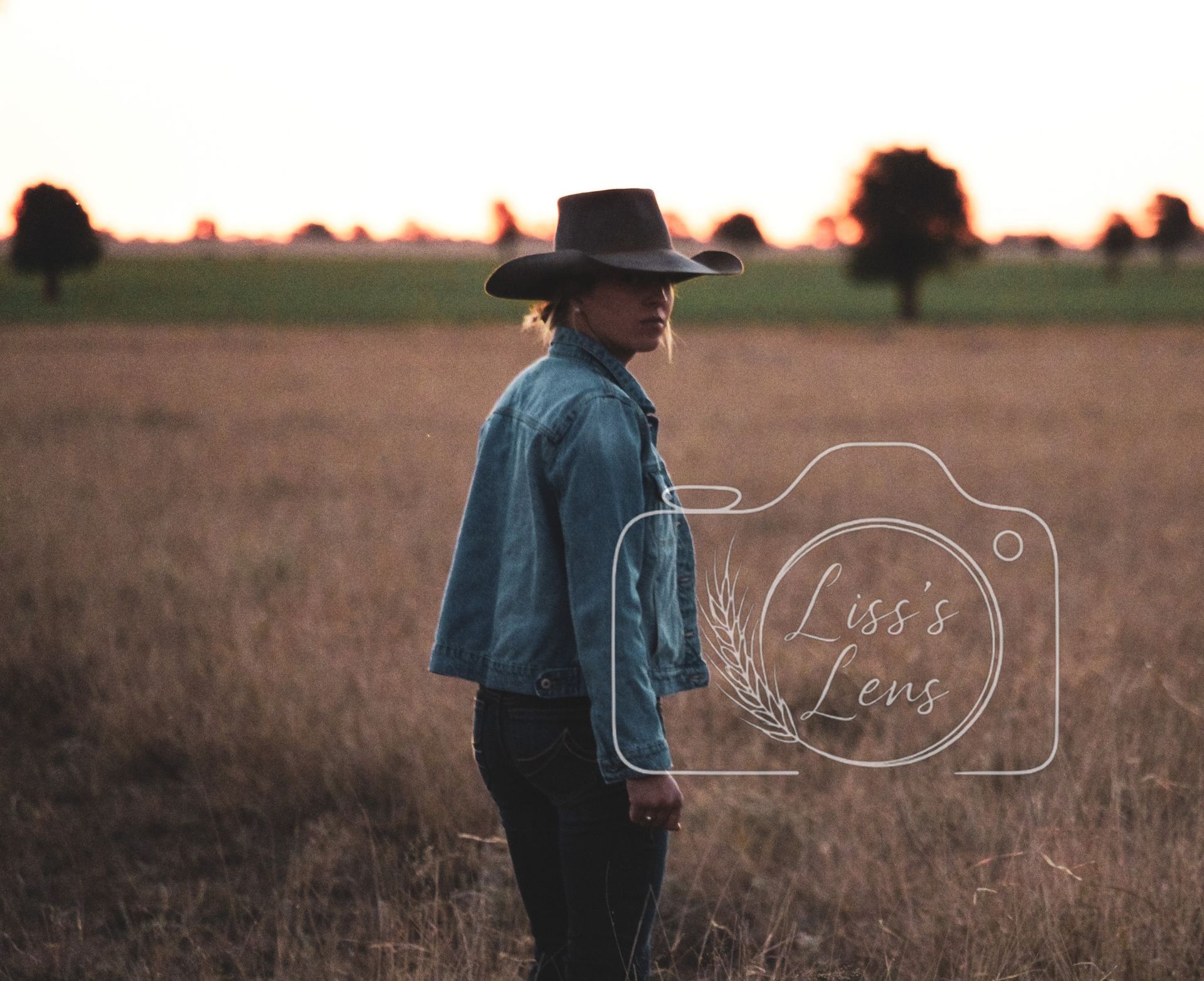 Woman in a cowboy hat and denim jacket stands in a field at sunset.