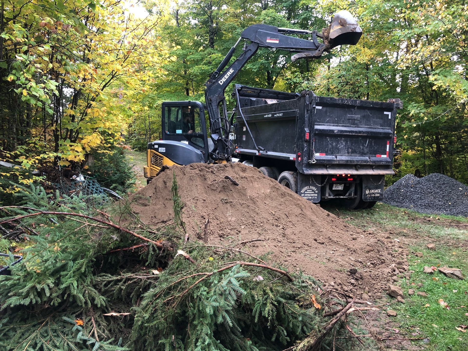 Une excavatrice chargeant de la terre dans la benne d'un camion à benne basculante, avec un tas de terre et de débris devant elle, dans une zone boisée.