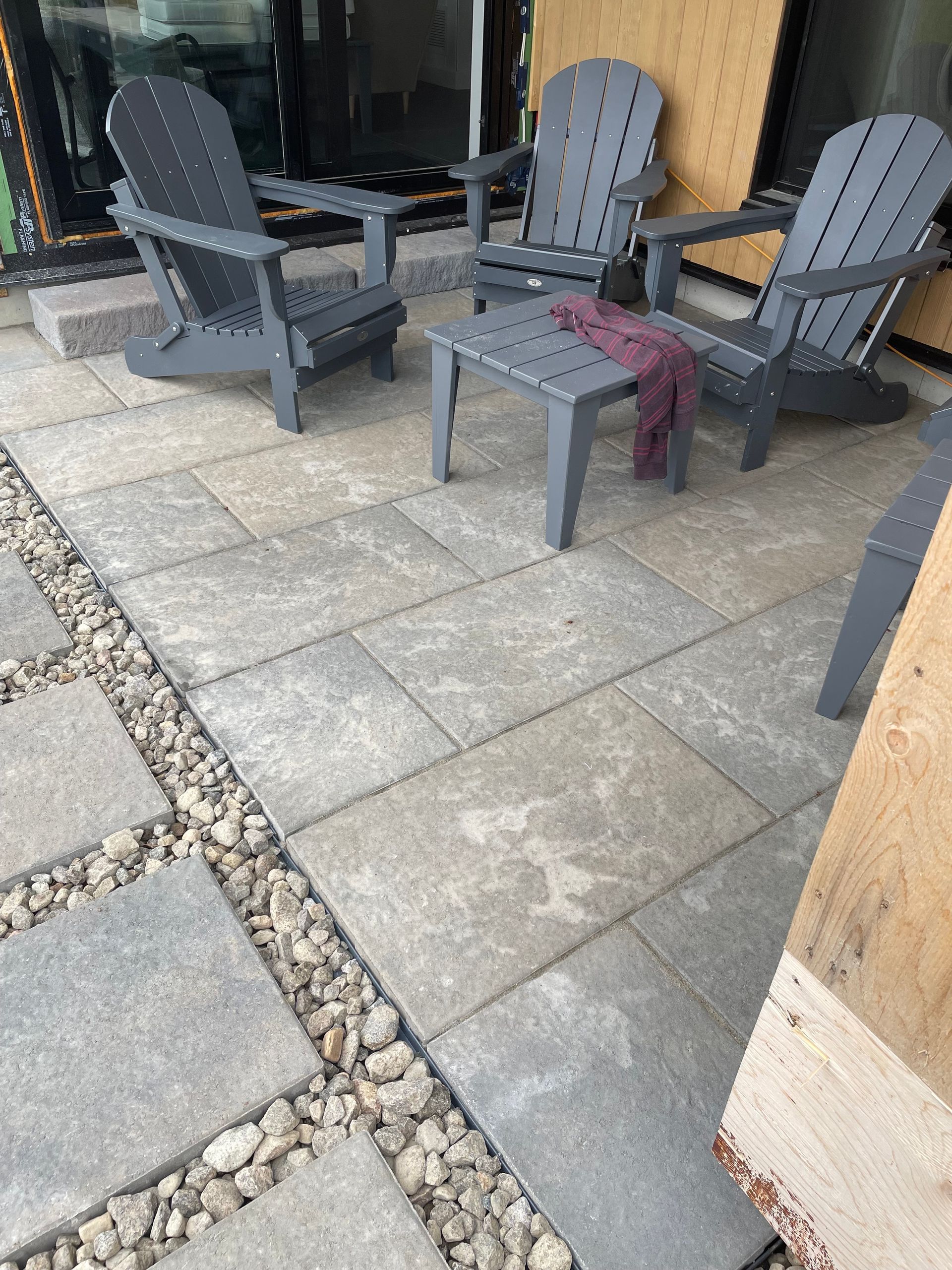 Patio avec chaises Adirondack grises et une petite table sur des pavés rectangulaires, bordé de gravier et de pavés carrés.