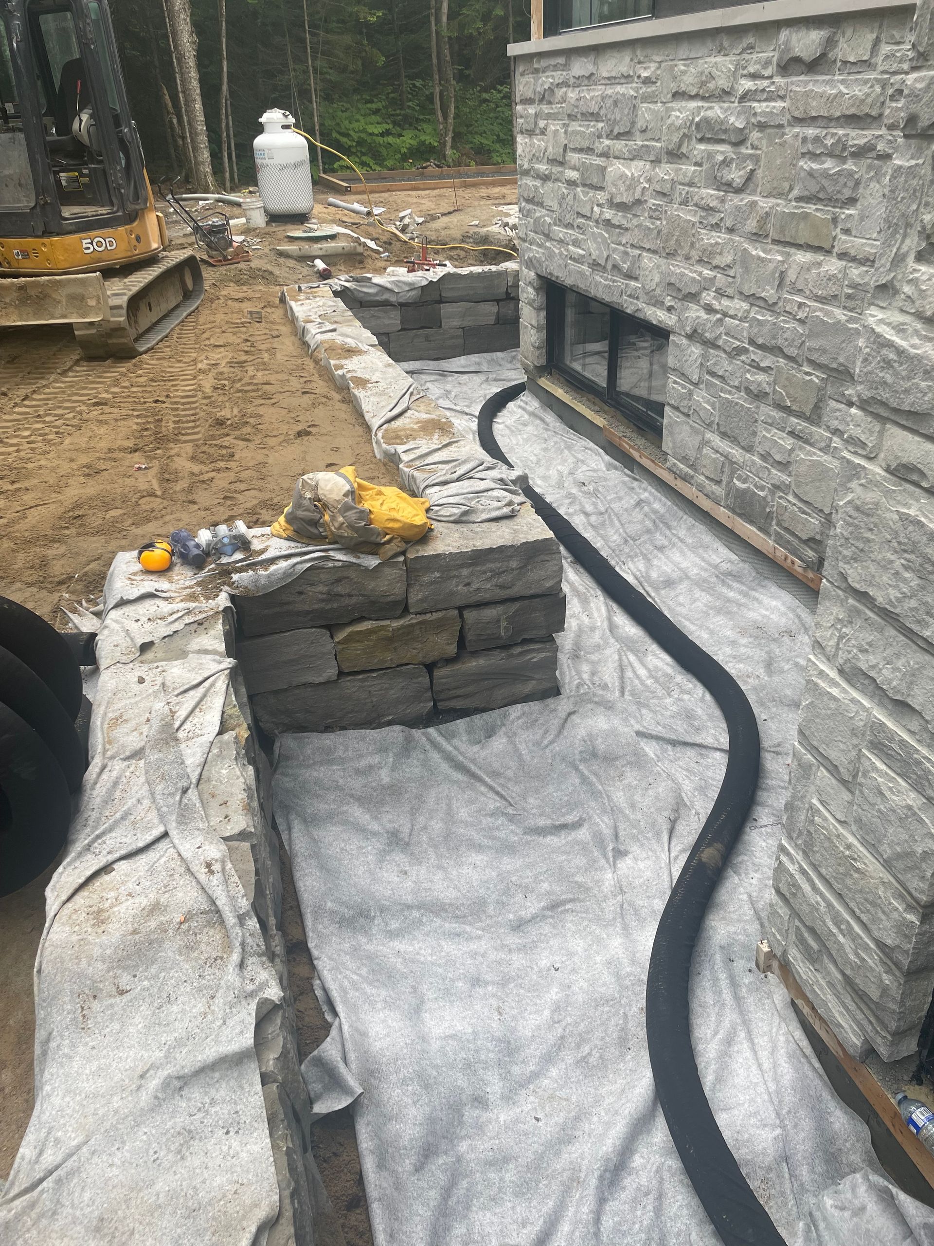 Chantier de construction avec mur de pierre en construction, tuyau de drainage noir posé sur un tissu gris et réservoir de propane en arrière-plan.