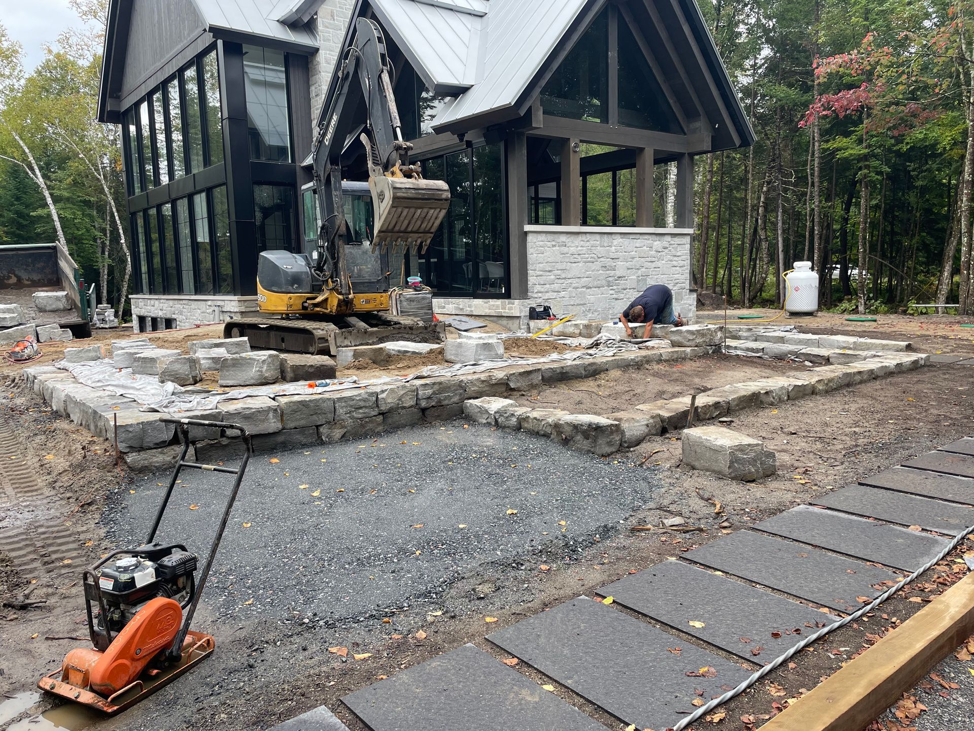 Chantier de construction avec une petite excavatrice, un ouvrier et un bâtiment avec de grandes fenêtres et une façade en pierre; gravier et pavés visibles.