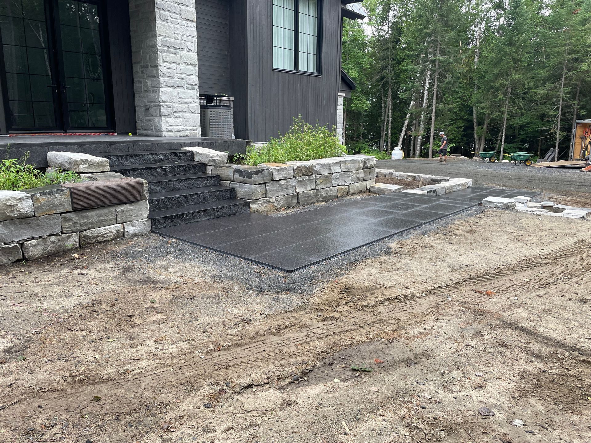 Une entrée de maison moderne avec des marches en pierre, un patio et un aménagement paysager gris foncé et un arrière-plan boisé.