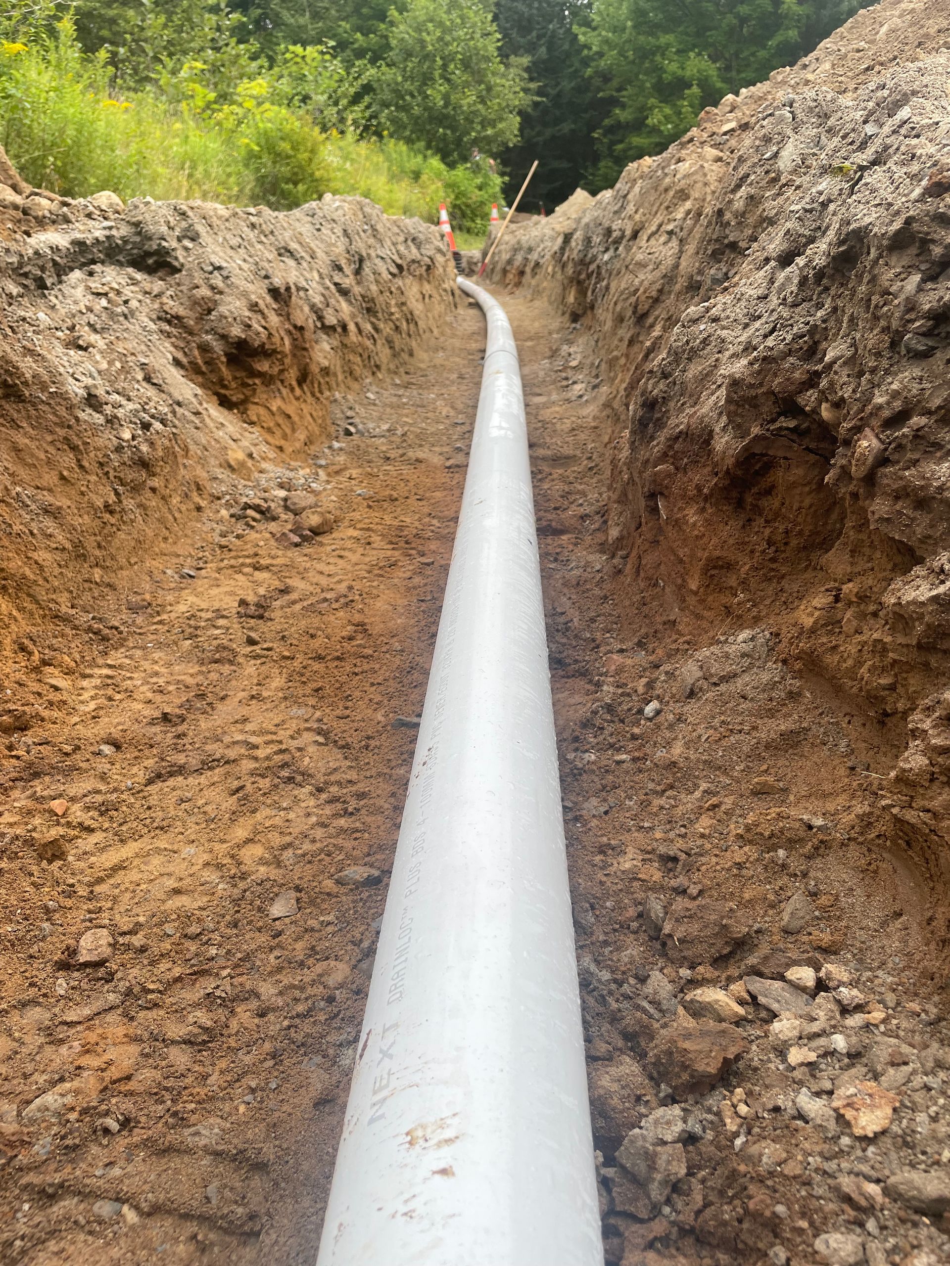 Tuyau blanc posé dans une tranchée longue et étroite dans de la terre brune, probablement pour un service public ou un drainage dans un environnement extérieur.