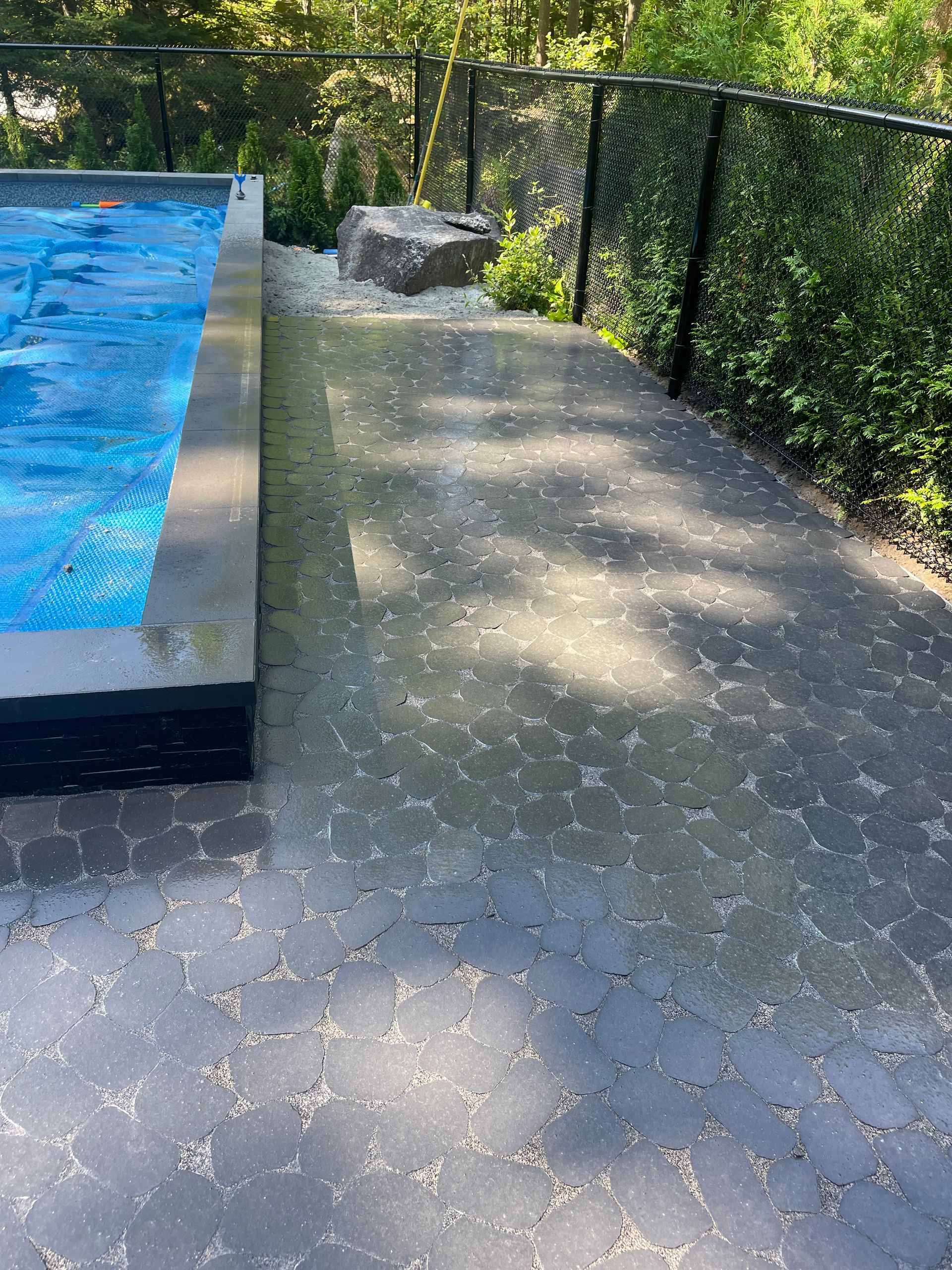 Une terrasse de piscine pavée de pierres sombres à motifs à côté d'une piscine recouverte d'une couverture bleue, entourée de verdure.