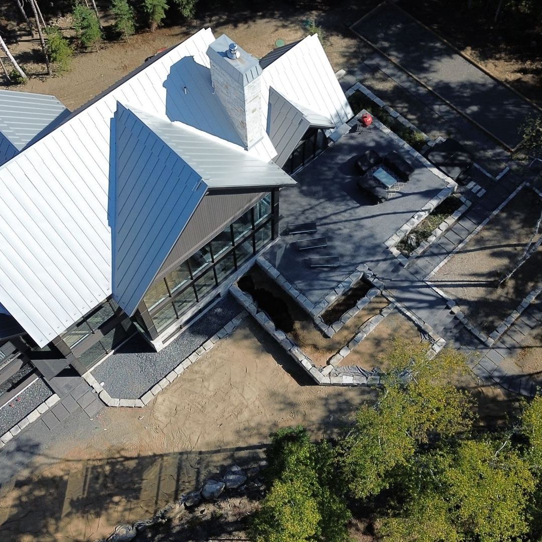 Vue en plongée d'une maison moderne avec un toit en métal blanc et de grandes fenêtres, située sur un patio en pierre avec une cheminée.