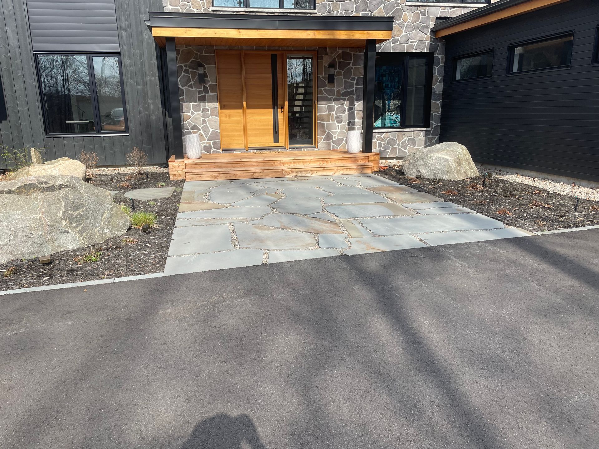 Entrée d'une maison moderne avec façade en pierre, porte en bois et une allée pavée menant à une allée en asphalte noir.