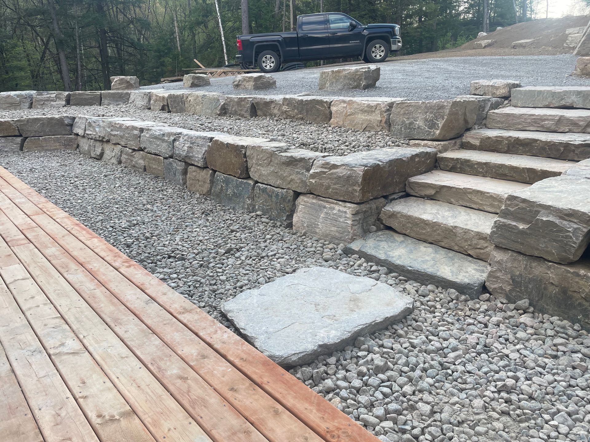 Mur de soutènement en pierre et marches menant à une allée de gravier avec une camionnette noire, à côté d'une terrasse en bois.