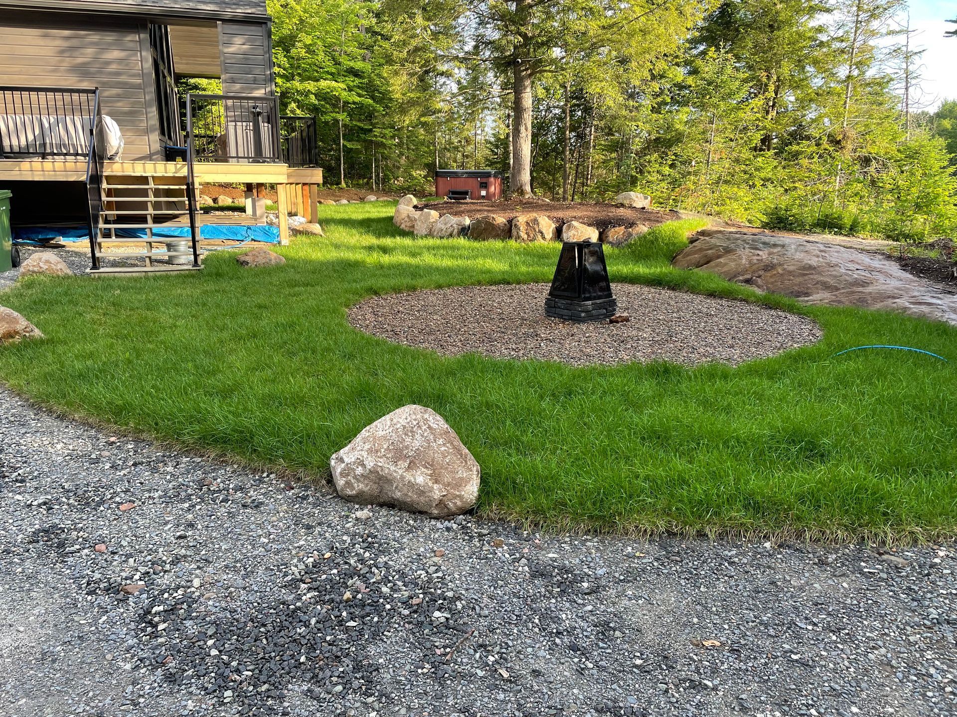 Un jardin avec un chemin de gravier, une pelouse verte, un foyer et des arbres. Un chalet avec terrasse se trouve à gauche.