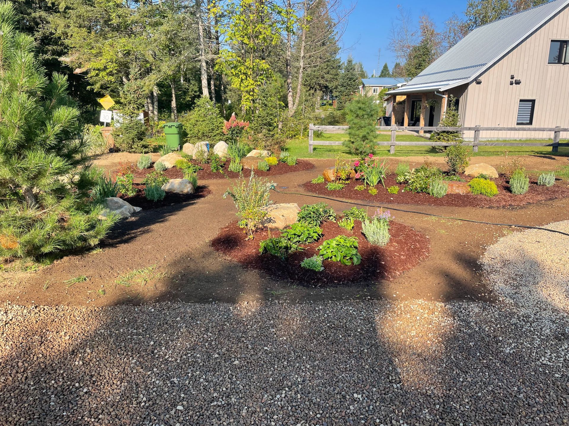 Un jardin paysager avec diverses plantes, des roches et du paillis devant un bâtiment avec un toit gris et une structure en bois.