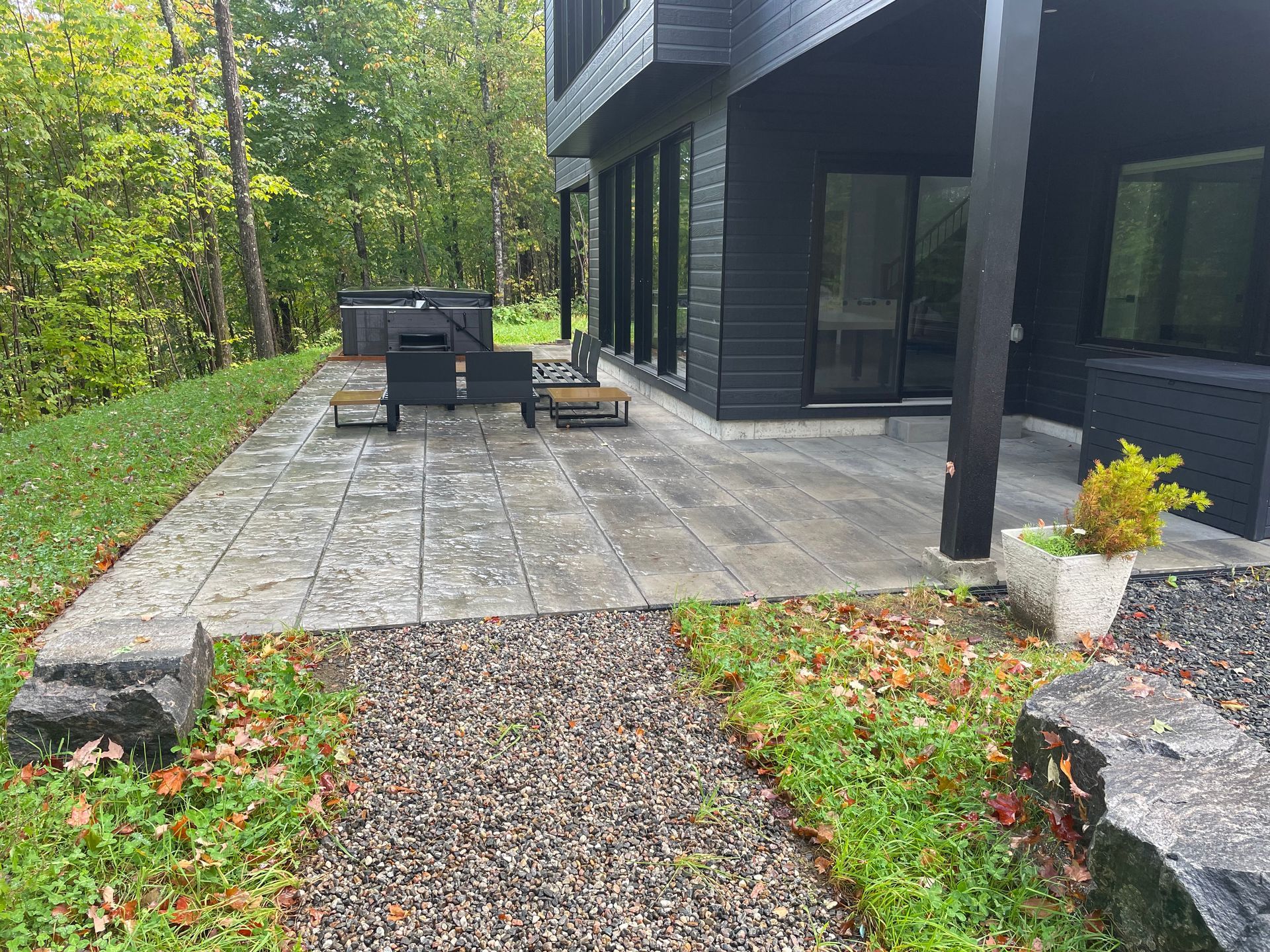 Une maison noire moderne avec un patio en pierre, des chaises longues et un jacuzzi dans un cadre boisé. Un chemin de gravier mène au patio.