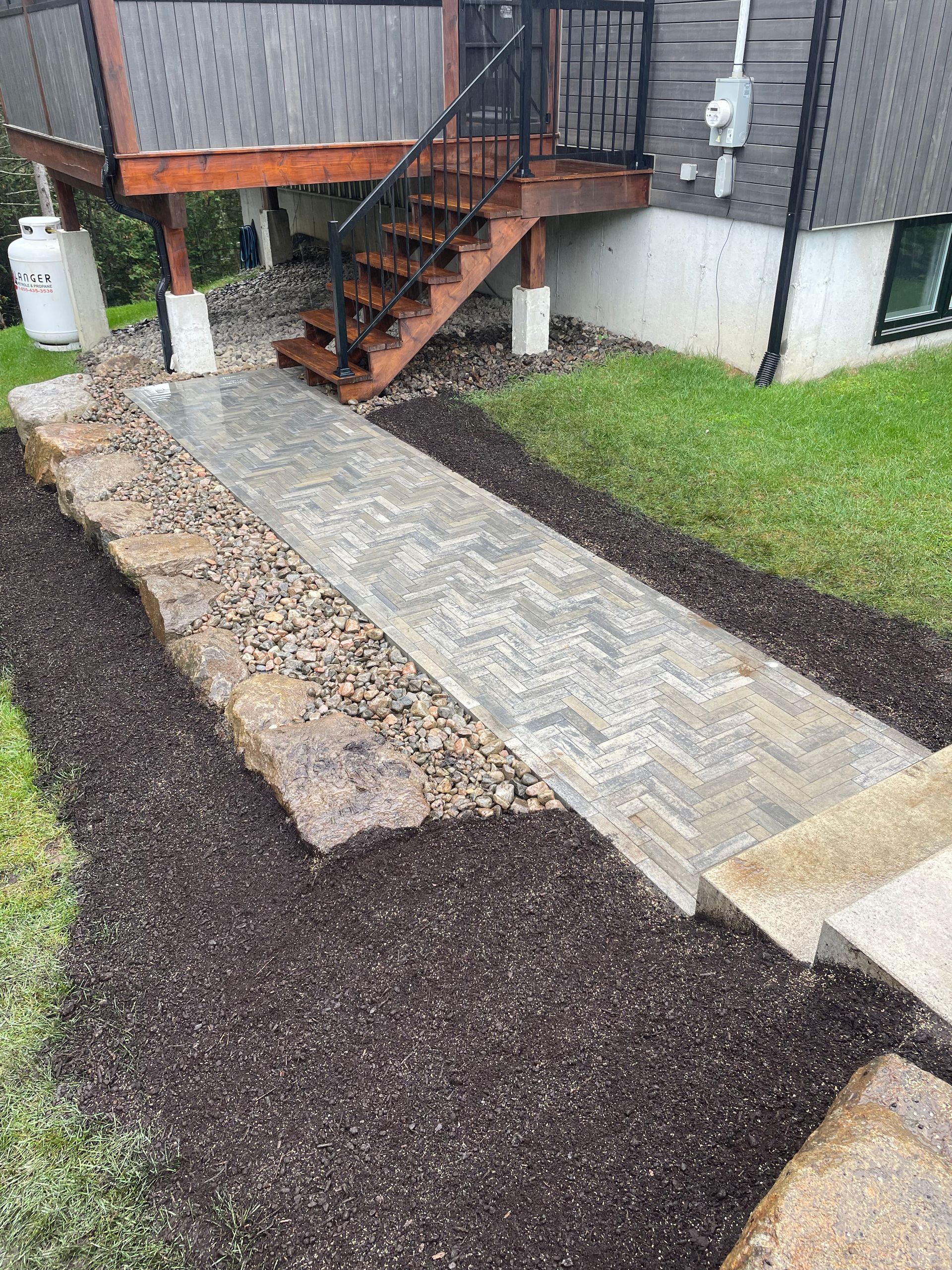 Un chemin en briques mène à une terrasse avec des escaliers, bordée de paillis et de pierres d'aménagement paysager sur une pelouse herbeuse.