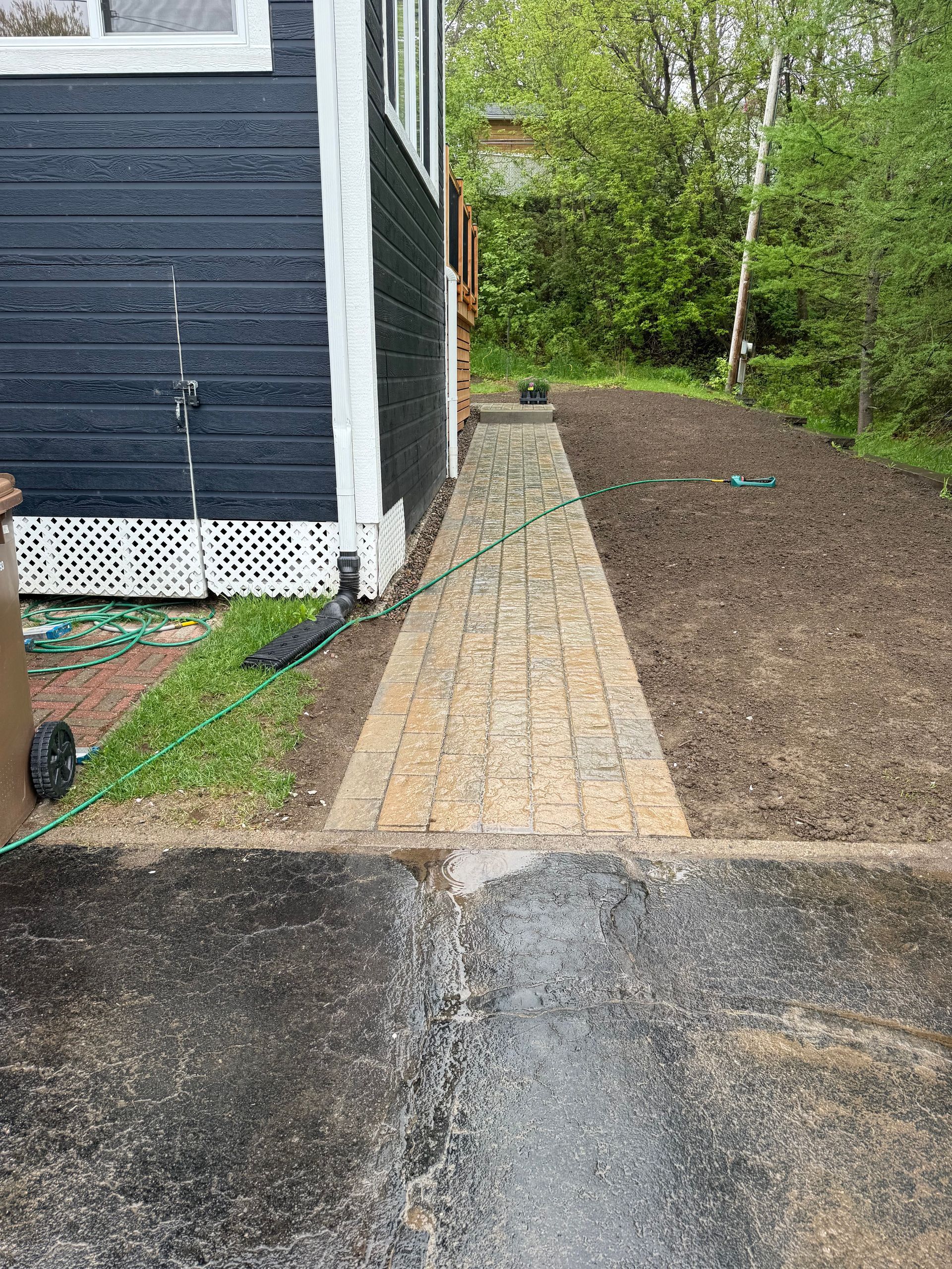 Un chemin de briques mène d'une zone pavée humide et sombre à un bâtiment. Il longe un jardin nu et une pelouse verdoyante.