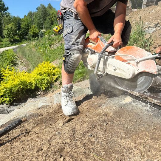 Un homme utilise une scie à béton à l'extérieur pour découper une surface grise. Il porte un short gris, des genouillères et des bottes de travail ; l'eau gicle.