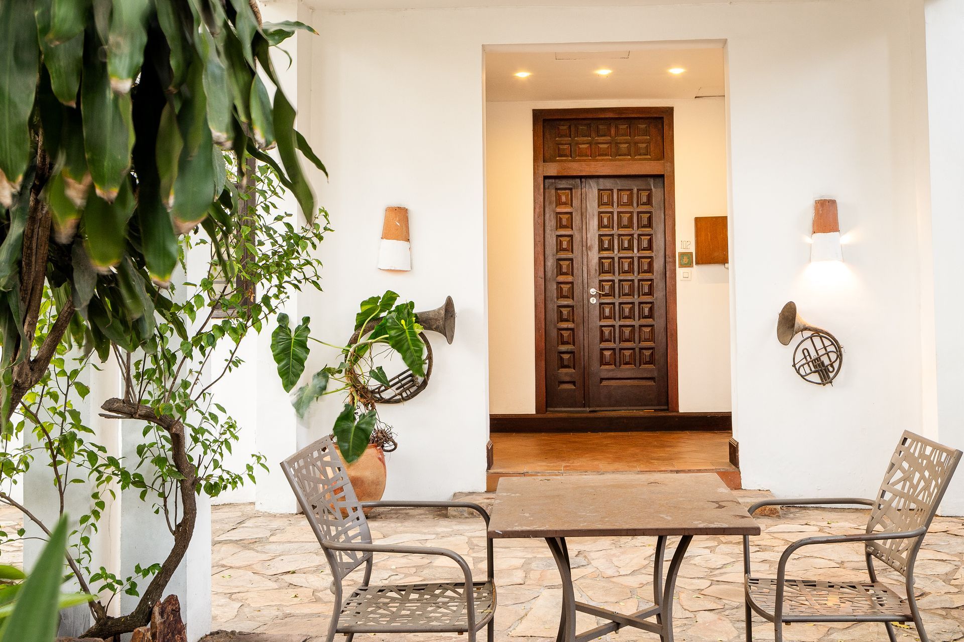A patio with a table, two chairs, and potted plants in front of an ornate wooden door in a white building.