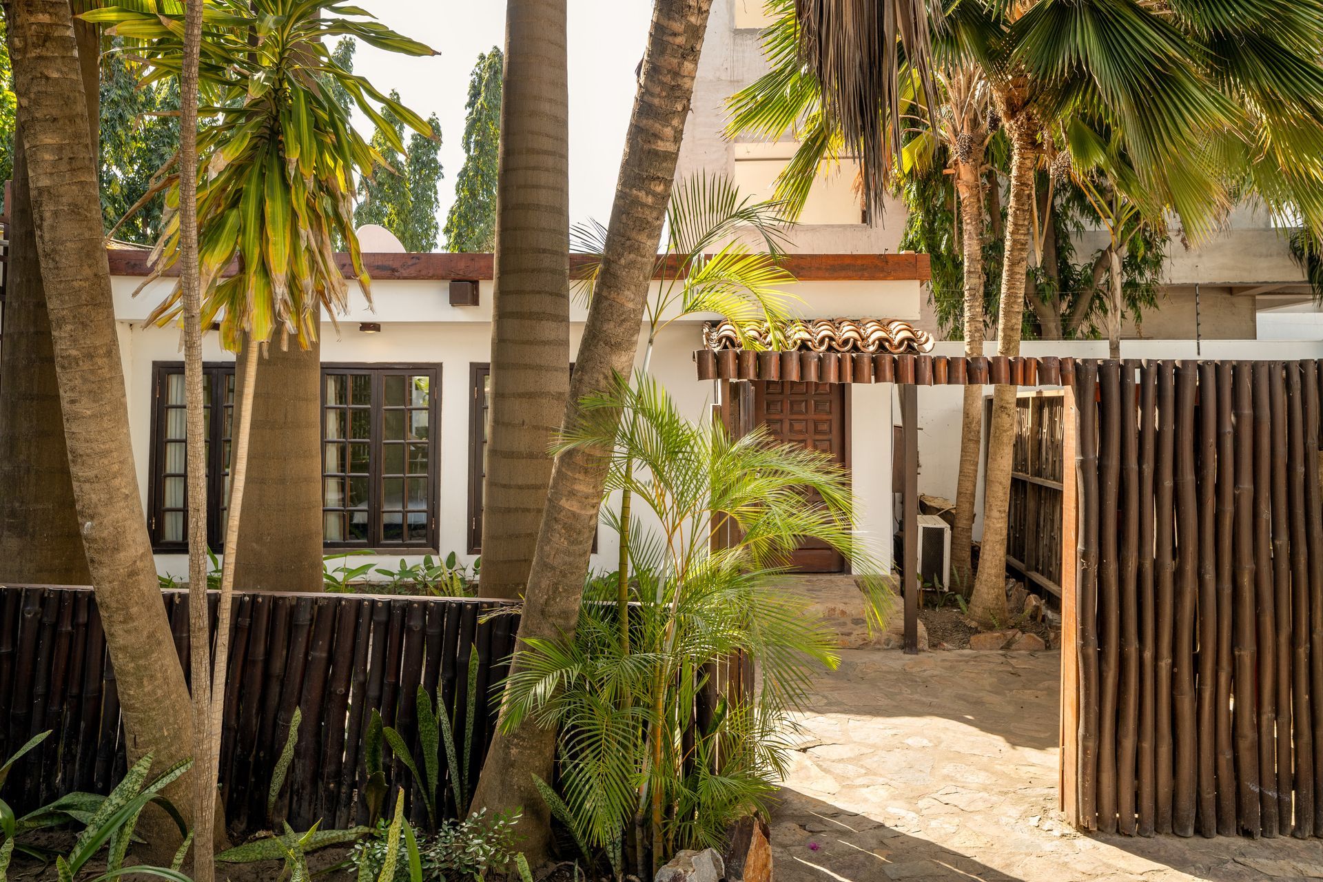 A white building with dark-framed windows behind a rustic wood-stick fence and palm trees, bathed in warm sunlight.