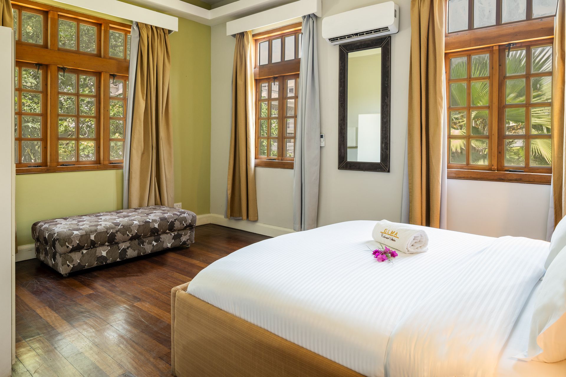 A hotel bedroom featuring a bed with white linens, a patterned bench, wood floors, and windows with gold curtains.