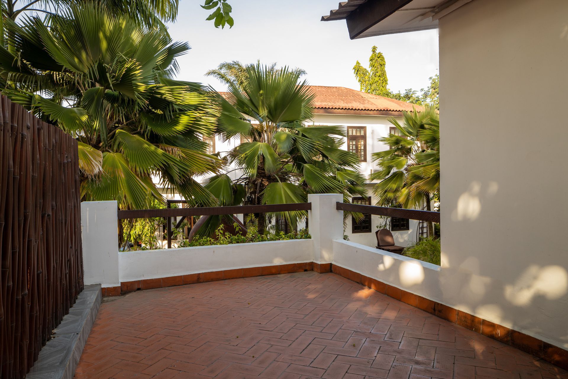 A sunny, brick-paved balcony with a brown railing, framed by lush palm trees and white building walls.