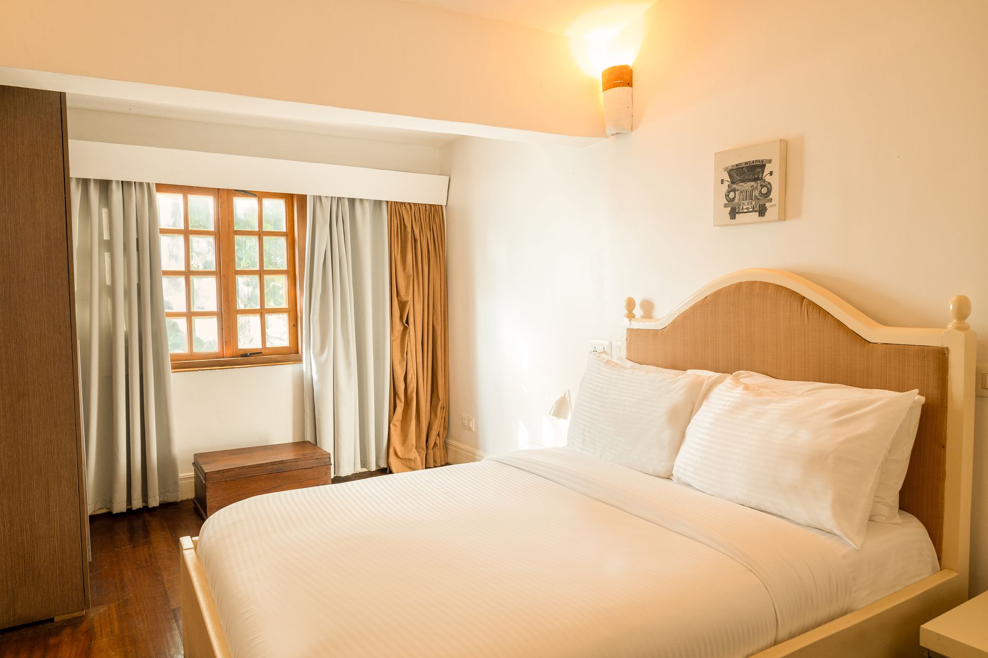A clean bedroom with a white-made bed, wooden floors, a tall wardrobe, and a window with light and dark curtains.
