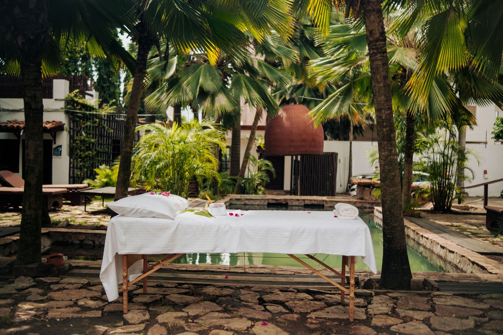 A white massage table stands outdoors on a stone patio by a pool, surrounded by lush palm trees.