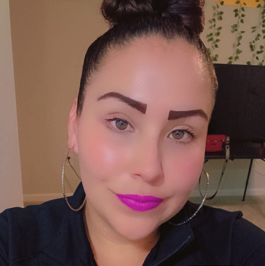 Woman in black uniform with logo, in a car, smiling at the camera. Brown eyes and eyebrows.