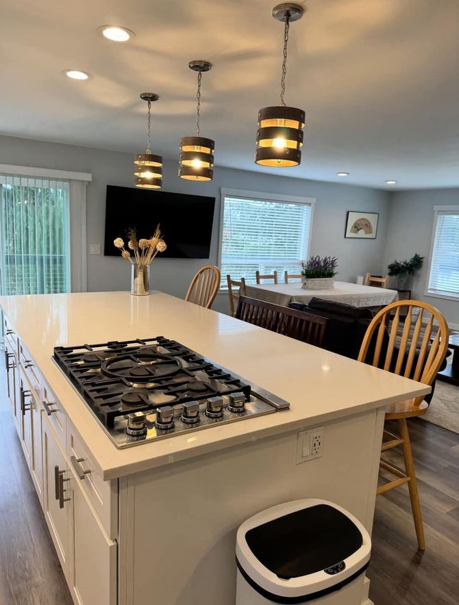 Modern kitchen with island, cooktop, pendant lights, and dining area. White cabinets, light gray walls, and wood floors.