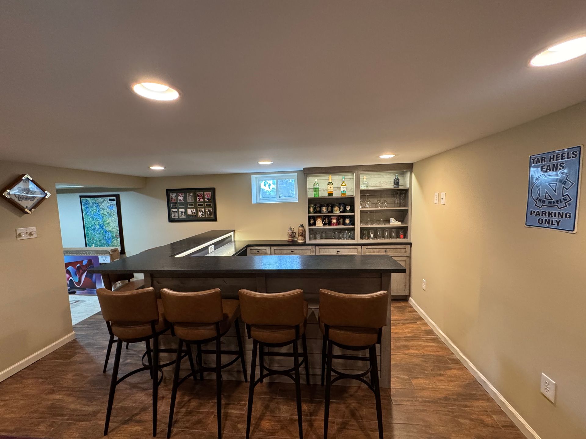 A finished basement with a dark wooden bar, four brown stools, recessed lighting, and shelving stocked with glassware.