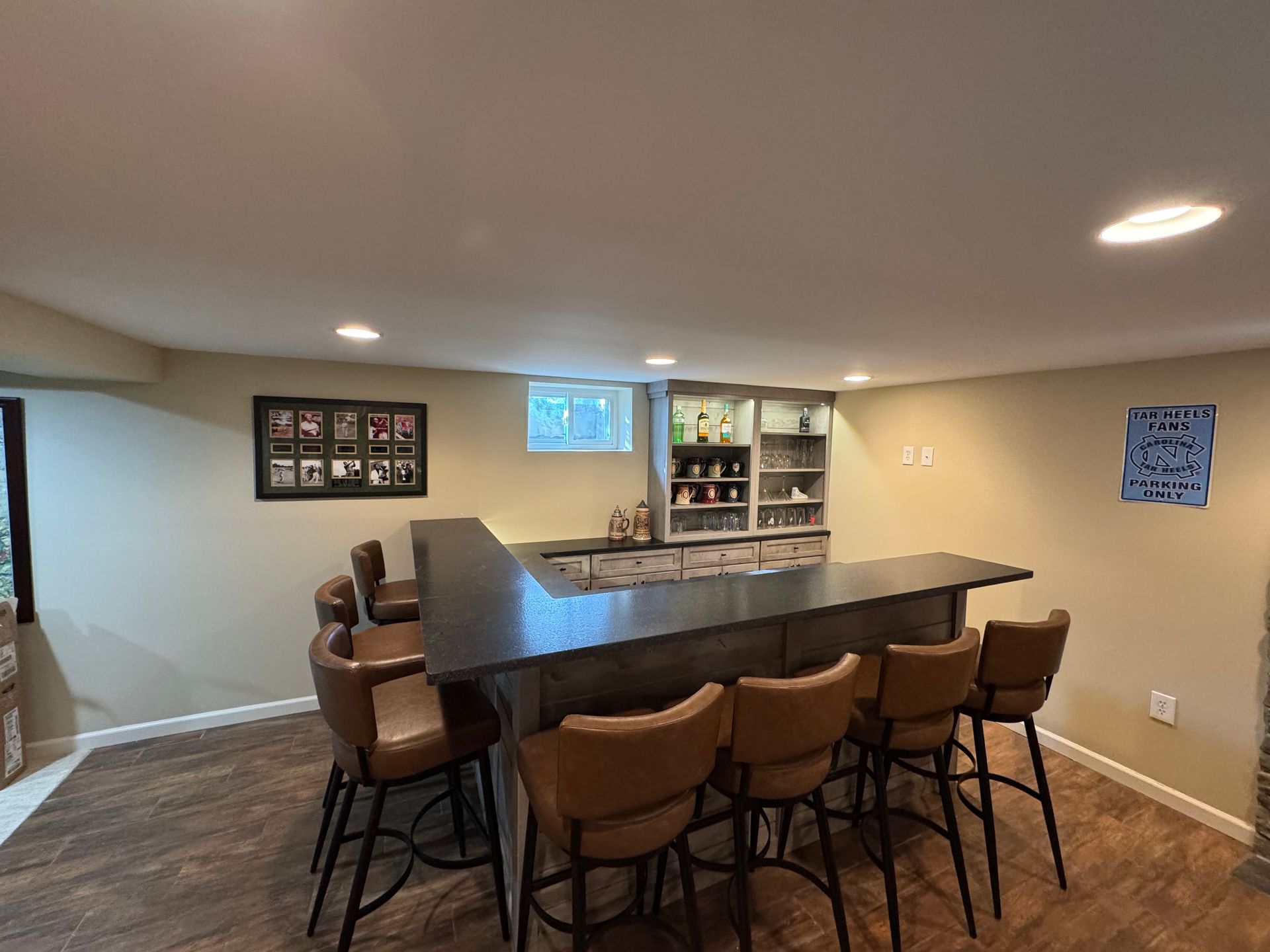 A home basement bar with dark countertops, wood flooring, and five tan leather swivel stools, plus a back display shelf.
