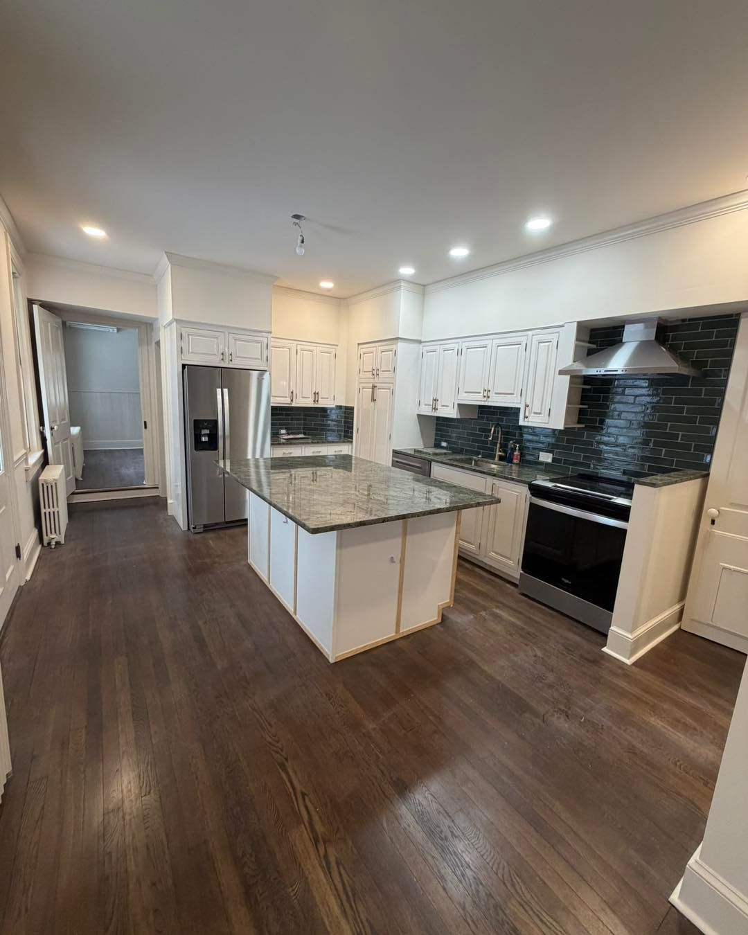 A bright, modern kitchen featuring white cabinets, a granite-topped island, dark hardwood floors, and stainless appliances.