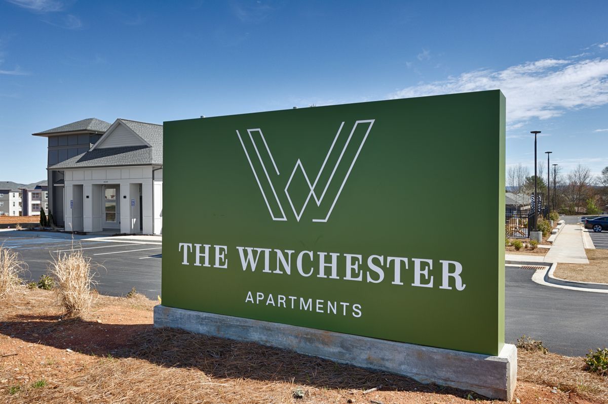 Sign for The Winchester Apartments with logo on green background, building in background.