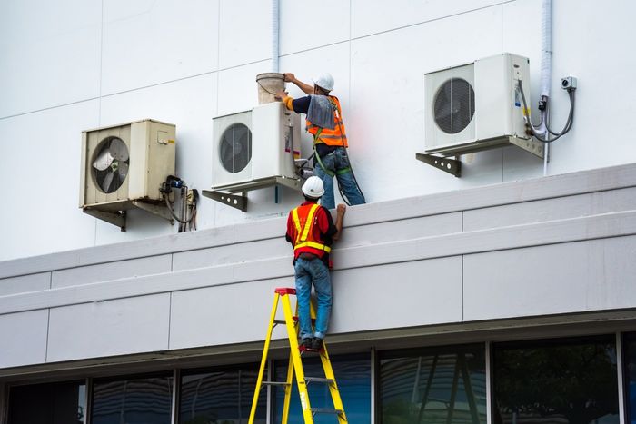 Due operai installano unità di condizionamento sul tetto di un edificio industriale, uno dei quali su una scala.