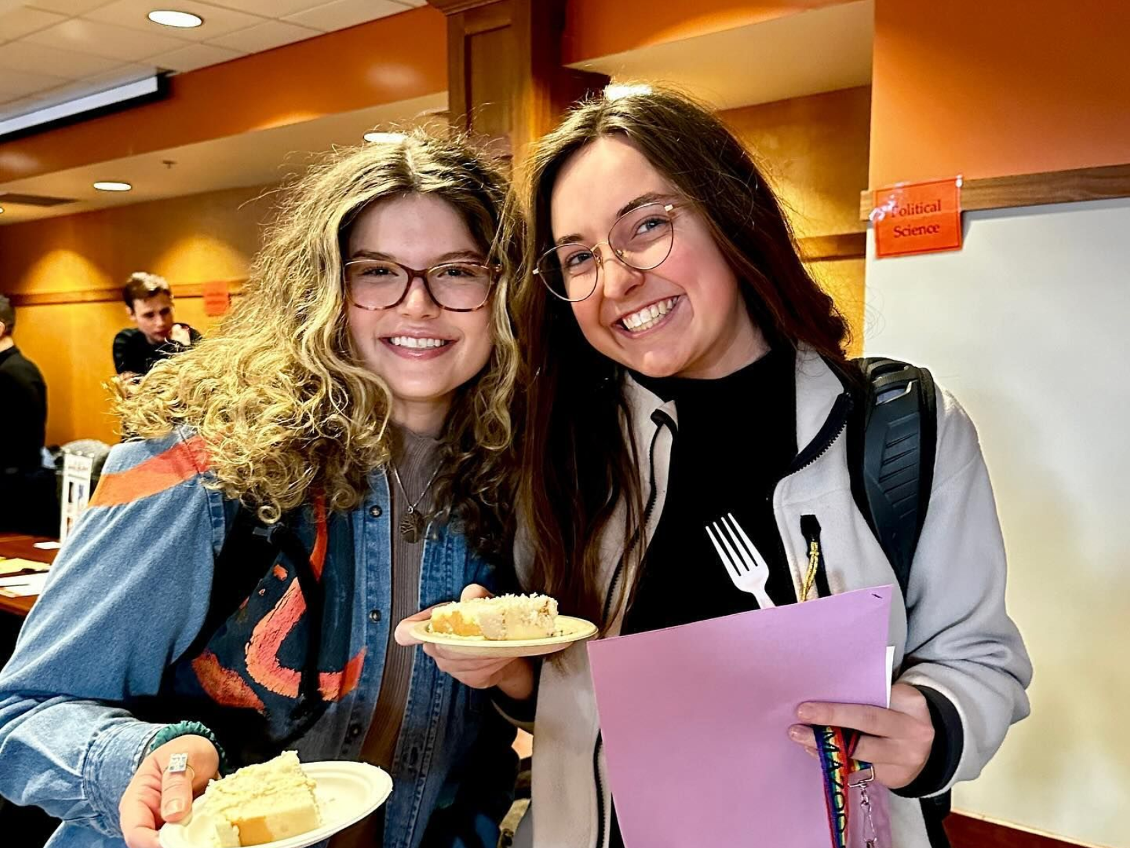 Two smiling people with plates of cake, one with a fork in their shirt, indoors.