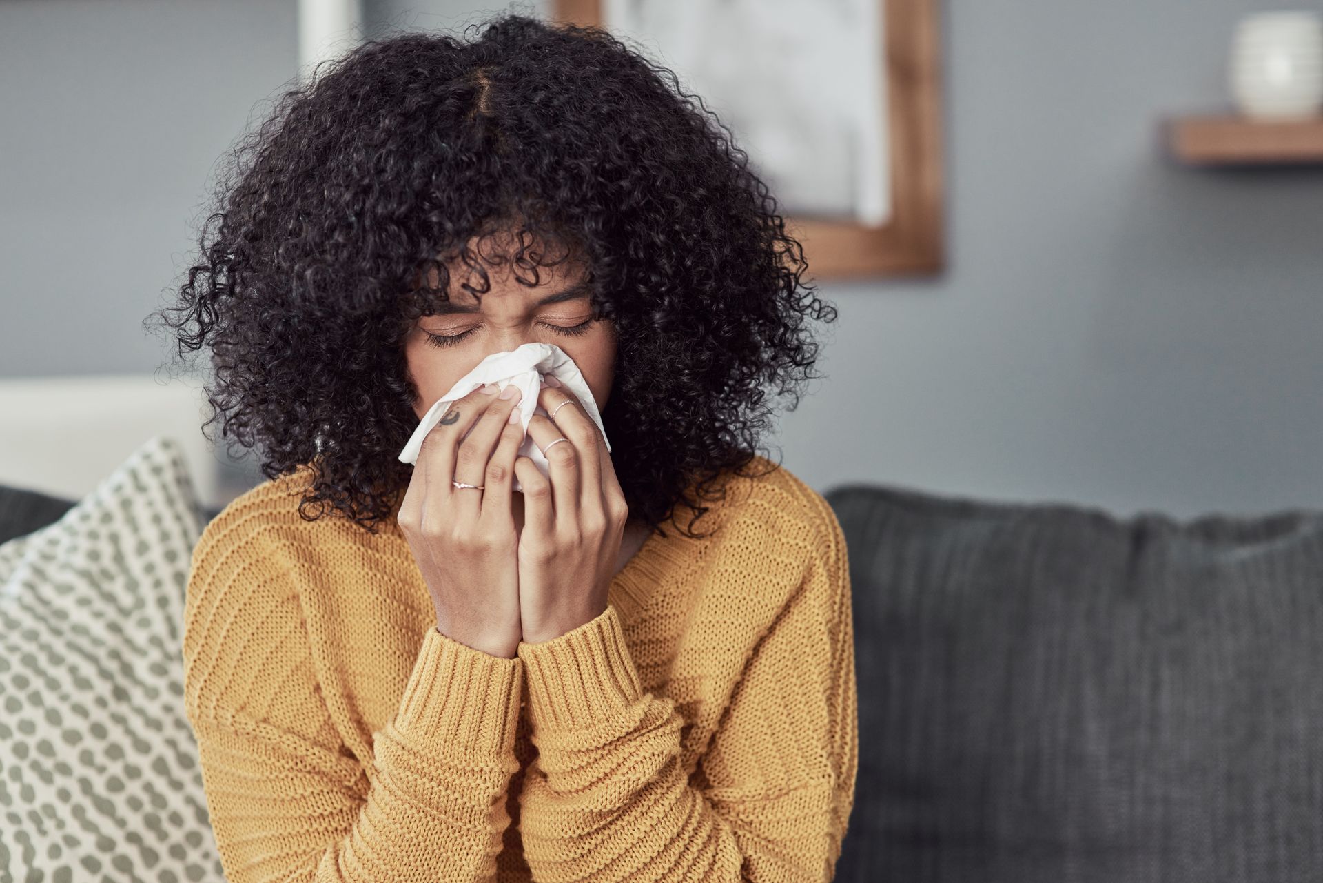 A person with dark, curly hair wearing a yellow sweater sits on a couch, holding a tissue to their nose.