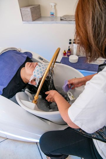 A professional washing a person's hair in a salon sink, with the client wearing a protective face shield.