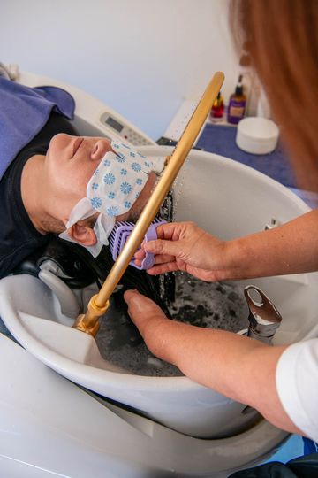 A stylist uses a specialized water spray tool and brush to wash a client's hair in a salon sink.