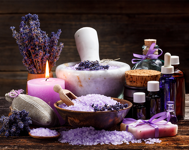 A wooden table topped with bowls of lavender salt , candles , essential oils and soap.
