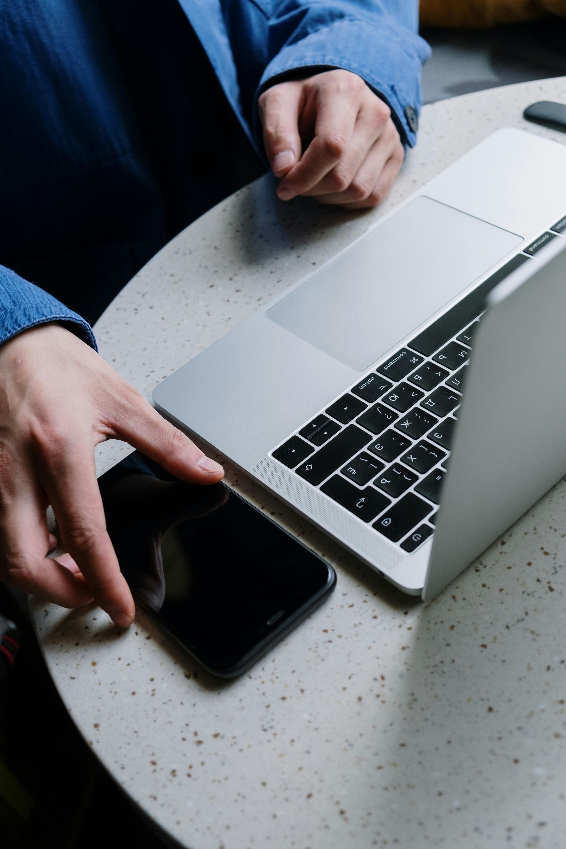 Person using a laptop and a smartphone on a speckled white table. Blue shirt sleeves are visible.